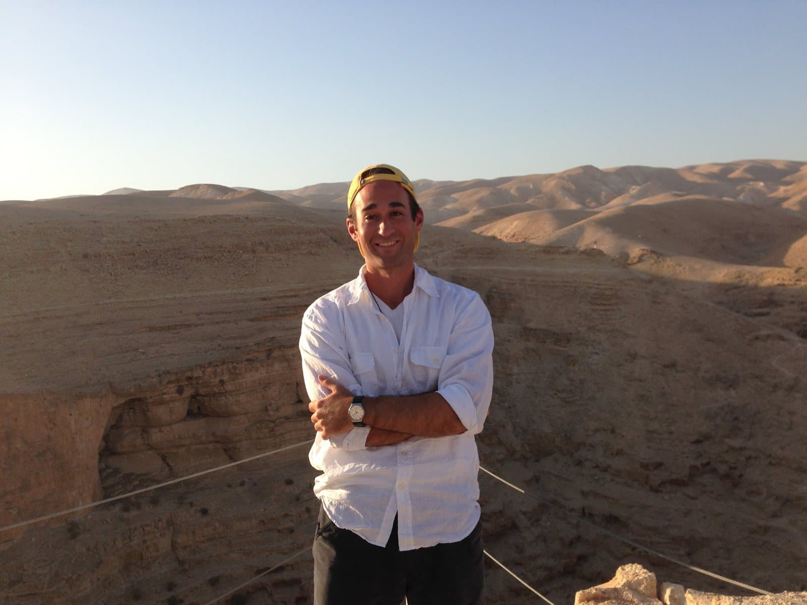 James Platania standing with arms folded at the edge of a canyon in the Judean Desert, in a white shirt and a yellow cap worn backwards, bare hills stretching to the horizon behind him.